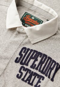 Camisa polo gris con cuello blanco, con un gran texto bordado en azul marino que dice "SUPERDRY STATE". Etiqueta visible dentro del cuello. La textura es suave.