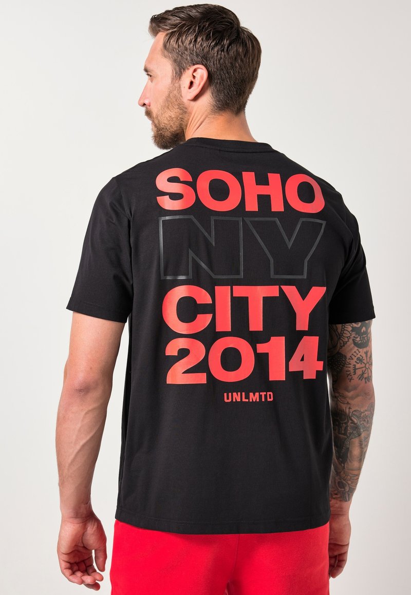 Hombre con barba y brazo derecho tatuado, vistiendo una camiseta negra con el texto rojo "SOHO CITY 2014" y "NY" contorneado en la parte de atrás, combinada con pantalones cortos rojos.