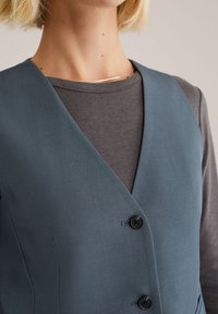 Vest teal cu o textură uniformă, având decolteu în V, nasturi negri și o croială ajustată, purtat peste o cămașă cu mânecă lungă de culoare gri închis.