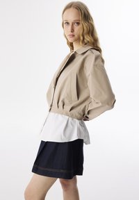 Helle beige Jacke mit einem taillierten Design, ausgestattet mit großen Taschen und einem weißen, geschichteten Saum, kombiniert mit einem dunklen Jeansrock und goldfarbener Naht.