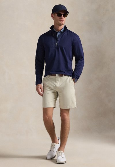 Μπλούζα με φερμουάρ μέχρι το στήθος στο χρώμα του ναυτικού, ανοιχτόχρωμα μπεζ σορτς, λευκά αθλητικά παπούτσια και καπέλο στο χρώμα του ναυτικού. Αξεσουάρ περιλαμβάνουν γυαλιά ηλίου και ρολόι. Ομαλή υφή υφάσματος.