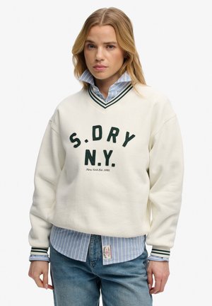 Kvinde iført en hvid S. DRY N.Y. sweatshirt over en blåstribet skjorte med krave og blå jeans, stående mod en hvid baggrund.