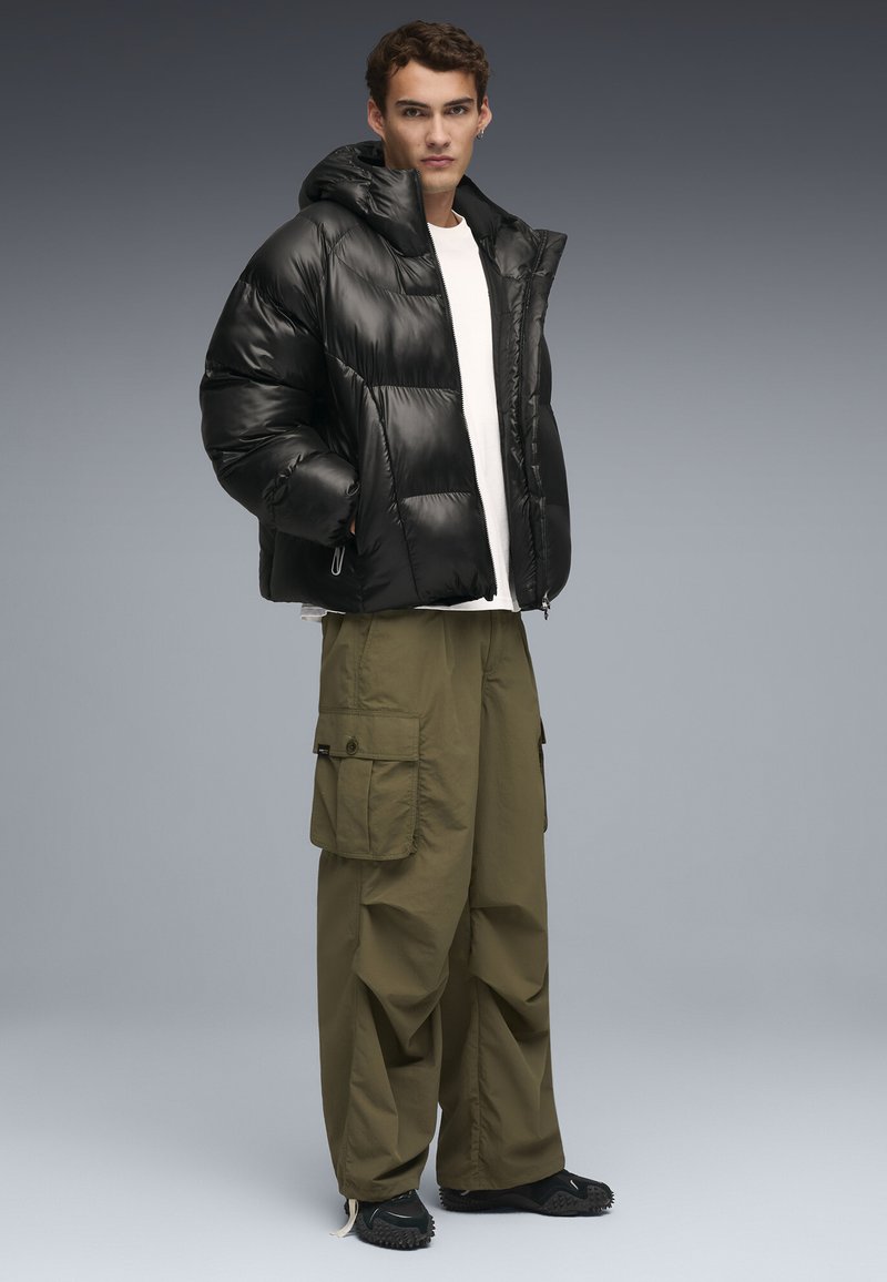 Giacca a vento nera con cappuccio, caratterizzata da una finitura lucida e un design trapuntato. Abbinata a pantaloni cargo verde oliva e calzature nere.