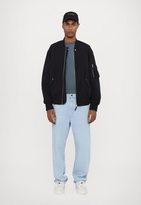 Black bomber jacket with a zip, light blue t-shirt underneath, light blue jeans, and white sneakers; a black cap completes the outfit.