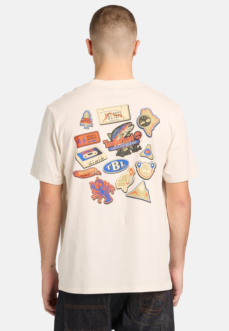 Homme portant un t-shirt beige avec des écussons colorés sur le thème de la nature et du vintage imprimés dans le dos, incluant un poisson, une cassette, un crabe et des montagnes.