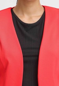 Röd lättviktsblazer med öppen front, lager-på-lager över en svart ribbad topp. Närbild visar den texturala kontrasten mellan tygerna.