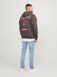 Άνδρας φορώντας σκούφο με κουκούλα σε σκούρο χρώμα με κόκκινη ροζέτα και κείμενο "Άγρια Νεότητα" στην πλάτη, ανοιχτόχρωμα μπλε τζιν και λευκά αθλητικά παπούτσια, κοιτάζοντας πλαγίως.