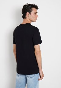 T-shirt en coton noir à manches courtes et col rond, porté par un modèle sur fond blanc uni, associé à un jean bleu clair.