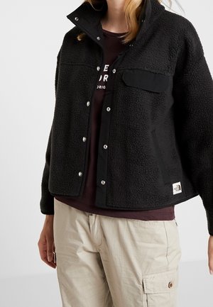 Person trägt eine schwarze Fleecejacke mit Druckknöpfen über einem dunklen Hemd und beigen Cargohosen, steht vor einem einfarbigen Hintergrund.