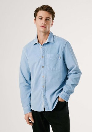 Chemise à manches longues bleu clair en tissu doux, avec une patte de boutonnage, une poche poitrine unique et des poignets boutonnés. Pantalons noirs.