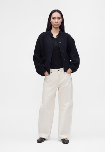 Femme aux longs cheveux noirs porte une veste noire boutonnée, un haut noir, un pantalon blanc large et des ballerines noires, debout les mains dans les poches.