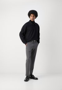 Črna polo majica z dolgimi rokavi v kombinaciji s sivimi krojenimi hlačami, dopolnjena s črnimi čipkastimi čevlji in črtastim pasom. Gladke teksture tkanin.