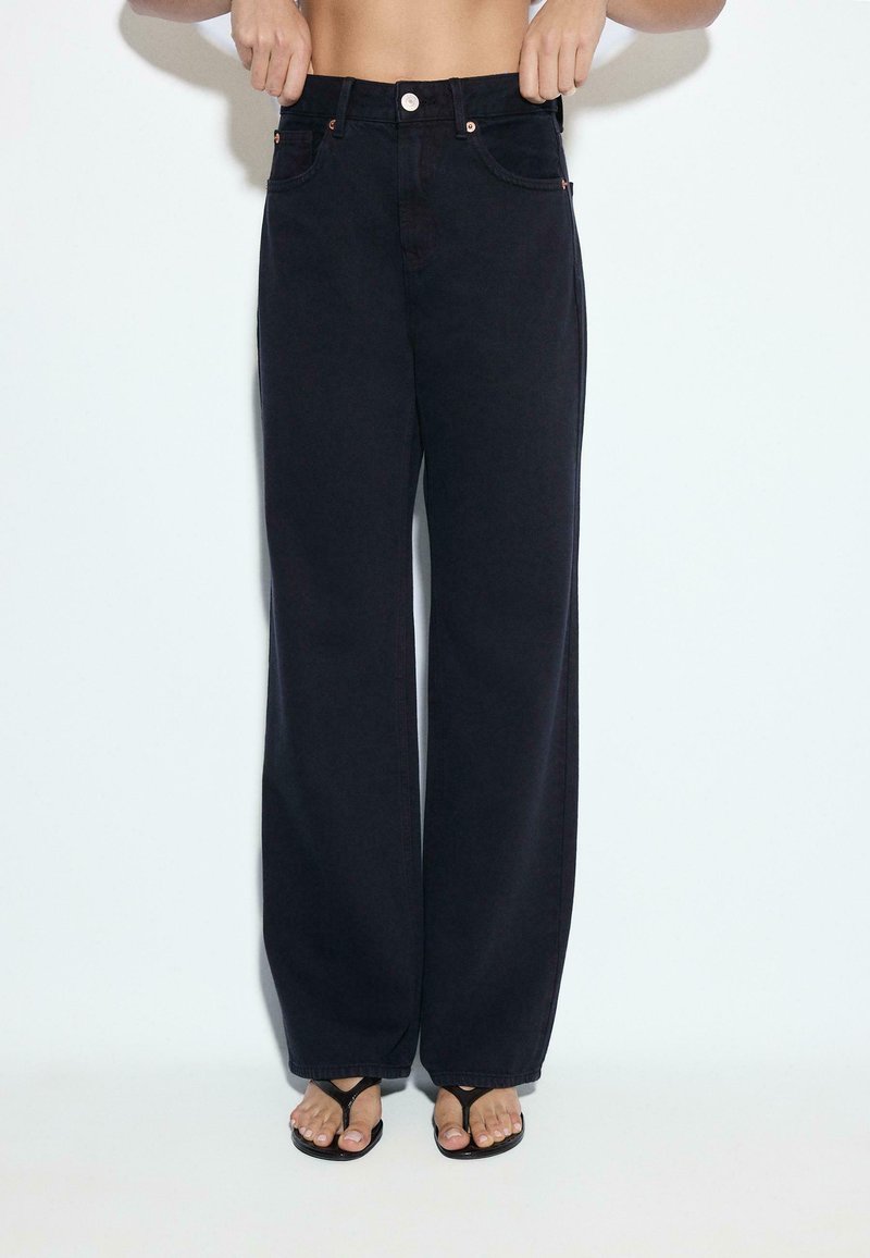 Personne portant un jean noir taille haute à jambes larges avec bouton et fermeture éclair, associé à des sandales noires à bouts ouverts sur un fond uni.