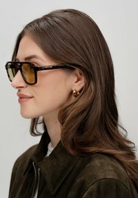 Lunettes de soleil en écaille de tortue avec des verres teintés bruns, monture rectangulaire et détails dorés sur les branches, portées avec une veste en daim noire.