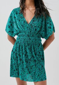Robe courte teal avec un décolleté en V, manches dolman courtes, taille froncée et un motif texturé noir sur l'ensemble.