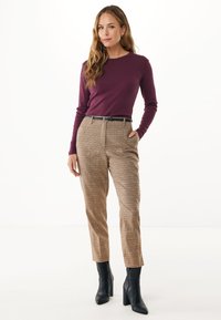 Eine Frau mit langen, welligen Haaren trägt ein enges, pflaumenfarbenes Langarmoberteil, beigefarbene karierte Hosen mit schwarzem Gürtel und schwarze, hochhackige Stiefeletten und steht.