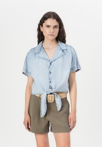 Ανοιχτό μπλε τζιν πουκάμισο κοντομάνικο δεμένο στη μέση, σε συνδυασμό με ελαιόχαρτες σορτς και ένα μπεζ ζωνάκι με τετράγωνη αγκράφα.