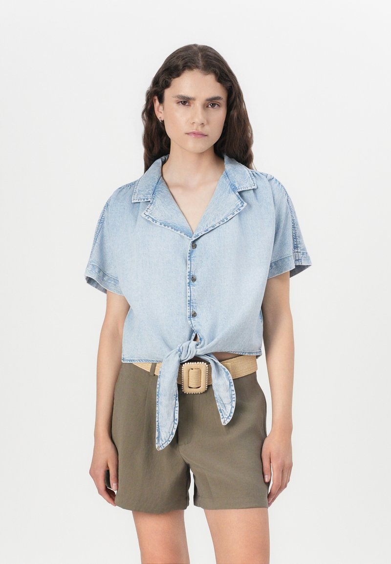 Light blue denim short-sleeve shirt tied at the waist, paired with olive green shorts, and a beige belt with a square buckle.