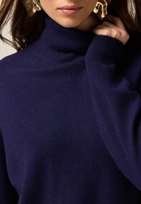 Navyblauer Rollkragenpullover aus einem weichen Material mit lockerer Passform und langen Ärmeln. Teilweise sichtbare goldene Statement-Ohrringe.