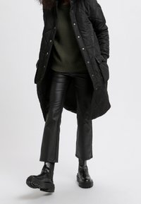 Abrigo largo negro acolchado, suéter de punto verde, pantalones acampanados de cuero sintético negro y botas negras gruesas. Diseño sencillo, tela texturizada.