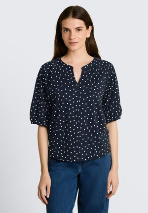 Jeune femme aux cheveux raides et foncés porte un chemisier bleu marine à pois blancs et un jean bleu, debout devant un fond uni.