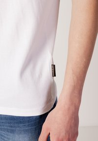 T-shirt en coton blanc avec une finition texturée, étiquette sur la couture latérale visible. Main reposant sur un jean en denim bleu. Design simple à col rond.