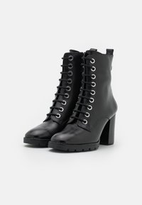 Un par de botas negras de cuero con cordones hasta el tobillo, con tacones gruesos y suelas de tracción, exhibidas sobre un fondo blanco.