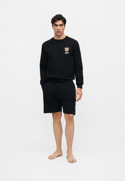 Άνδρας όρθιος ξυπόλυτος που φοράει μαύρο μακρυμάνικο πουκάμισο με λογότυπο αρκουδάκι και μαύρα σορτς μέχρι τα γόνατα σε απλό φόντο.