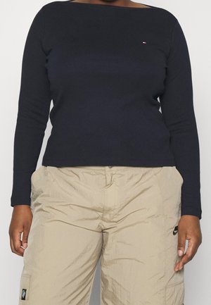 Osoba ubrana w czarną bluzkę z długim rękawem oraz beżowe spodnie typu cargo Nike, z rękami opartymi wzdłuż ciała.