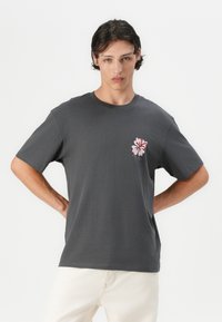 Grijze katoenen t-shirt met een roze bloemenontwerp op de linkerkant van de borst, korte mouwen en een klassieke ronde hals.