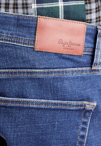 Plave traperice s smešnim kožnim umetkom označenim s "Pepe Jeans London." Imaju narančasti šav i teksturiranu završnu obradu.