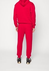 Červená mikina s kapucí a souprava tepláků vyrobená z fleecového materiálu, obsahuje klokaní kapsu a žebrované manžety. Nosené s černými teniskami.