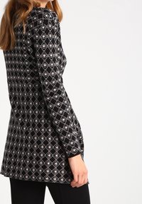 Kvinna iklädd en långärmad svart tröja med vita geometriska mönster, tillsammans med svarta byxor, visad från sidan mot en vit bakgrund.