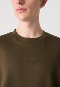 Sweatshirt à col rond vert olive fabriqué à partir d'un tissu lisse, avec un logo « Porsche BOSS » embossé au design minimaliste sur la poitrine.