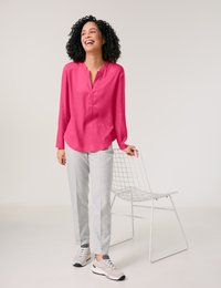 Bright pink blouse with a v-neck, light grey plaid trousers, and grey sneakers. The setting features a minimalist white wire chair.