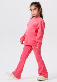 Jong meisje met lang, donker haar, gekleed in een bijpassende felroze sweatshirt en broek, die een zelfverzekerde pose aanneemt tegen een witte achtergrond.