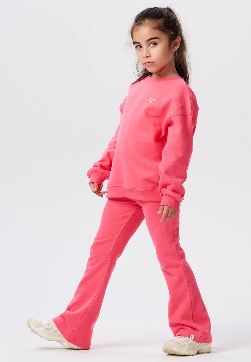 Jong meisje met lang, donker haar, gekleed in een bijpassende felroze sweatshirt en broek, die een zelfverzekerde pose aanneemt tegen een witte achtergrond.