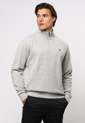 Grijze rits-sweater met een hoge kraag, geribbelde manchetten en een klein geborduurd logo op de borst, gedragen met donkere broek.