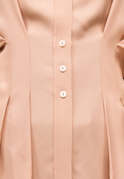 Κοντινό πλάνο εμπρόσθιου μέρους μπεζ πουκαμίσου από ύφασμα με κάθετες ραφές και τρία λευκά στρογγυλά κουμπιά που είναι κουμπωμένα στο κέντρο.