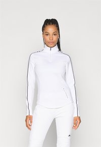 Top atletico bianco con zip al collo e maniche lunghe, caratterizzato da strisce laterali nere e una finitura texture lungo l'orlo e l'area della zip.