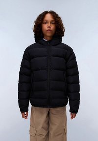 Criança com cabelo encaracolado, vestindo uma jaqueta acolchoada preta com fecho e calças cargo bege, de pé contra um fundo simples.