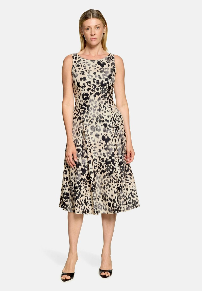 Femme portant une robe sans manches longueur genoux avec un imprimé léopard noir et beige et des talons ouverts noirs, debout devant un fond blanc.