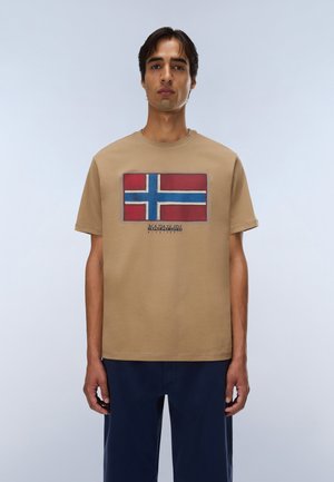 Hombre con camiseta beige con una bandera cruzada roja, blanca y azul y pantalones azul marino, de pie contra un fondo claro y liso.