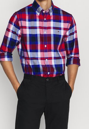 Homme portant une chemise à carreaux rouge, bleu, blanc et gris, à boutons, manches retroussées jusqu'aux coudes, et un pantalon noir, les mains dans les poches.