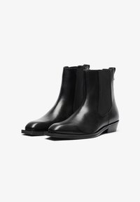 Botas de tobillo de cuero negro con paneles laterales elásticos, punta en forma de almendra y un tacón bajo apilado. Textura suave con un detalle de costura mínimo.