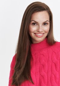 Jasnoróżowy sweter z dzianiny z wysokim, prążkowanym dekoltem, ozdobiony wzorami warkoczowymi. Długie, proste brązowe włosy i naturalny makijaż.