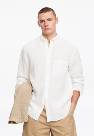 Hombre vistiendo camisa blanca de botones y pantalones cortos beige, sosteniendo un suéter de punto beige con la mano izquierda contra un fondo blanco.