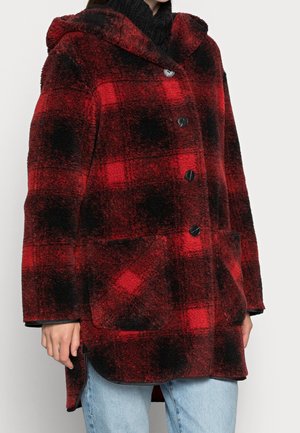 Vrouw die een rood-zwart geruit, zacht jasje draagt met grote knopen en zakken aan de voorkant, gecombineerd met lichtblauwe jeans.