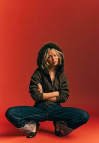 Kapuzensweater in bruin met bontaccenten, oversized donkerblauwe denim jeans met rafelige manchetten, en zwarte schoenen tegen een rode achtergrond.