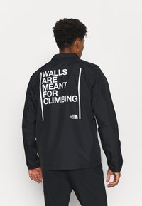 Homme portant une veste noire avec le texte blanc "Les murs sont faits pour être escaladés" et le logo The North Face dans le dos, se tenant devant un fond blanc.